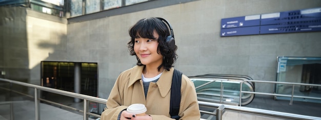 Portrait d'une jeune étudiante dans un casque boit du café dans la rue avec des trajets en sac à dos