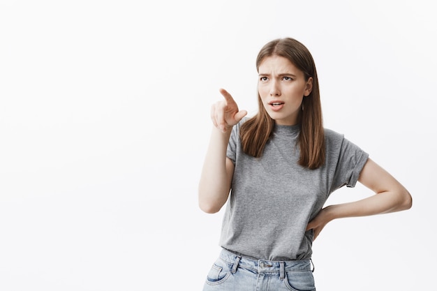 Photo gratuite portrait de jeune étudiante européenne confiante aux cheveux bruns dans des vêtements décontractés pointant sur la personne, regardant avec une expression faciale moyenne, se disputant avec une fille agressive au collège.