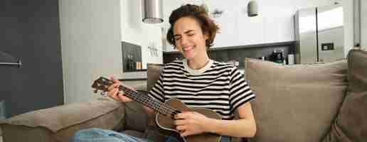 Photo gratuite portrait d'une jeune étudiante moderne jouant du ukulélé à la maison assise avec une petite guitare chantant et