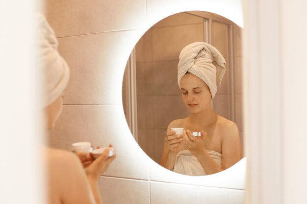 Portrait d'une jeune femme adulte debout dans la salle de bain devant un miroir avec de la crème dans les mains, faisant des soins de beauté à la maison après avoir pris une douche, enveloppée dans une serviette blanche.