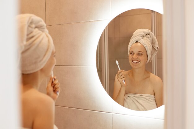 Portrait d'une jeune femme adulte souriante aux épaules nues se brossant les dents, posant dans la salle de bain après avoir pris une douche, debout avec une serviette blanche sur ses cheveux.