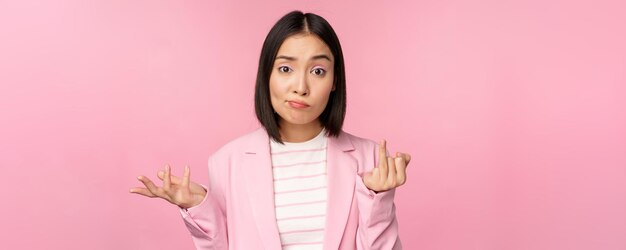 Portrait de jeune femme d'affaires asiatique vendeuse haussant les épaules et regardant confus désemparé de qch debout sur fond rose