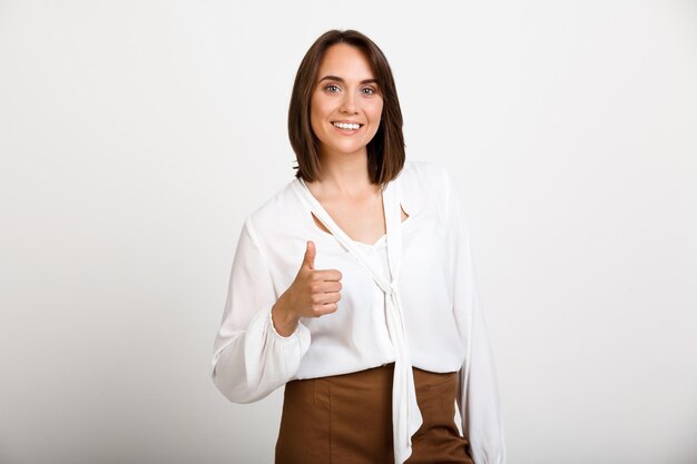 Portrait d'une jeune femme d'affaires prospère souriante, montrant le pouce vers le haut, regardant à l'avant, sur un mur blanc.