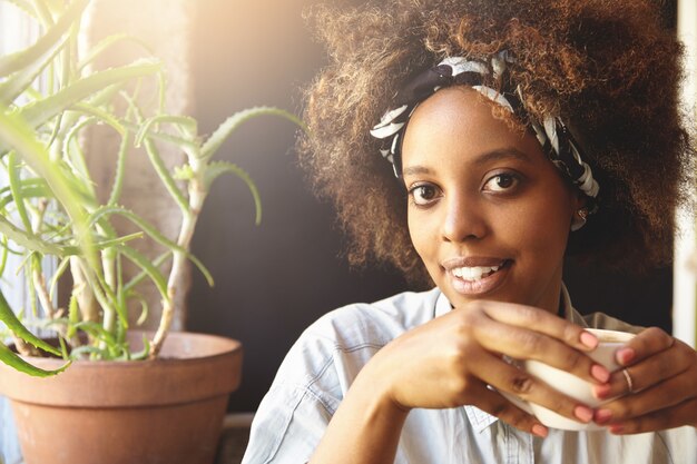 Portrait de jeune femme afro-américaine avec coupe de cheveux afro vêtue d'une chemise en jean élégante blanche, tenant une tasse de café chaud ou de thé avec un visage heureux, ayant du bon temps à la cafétéria