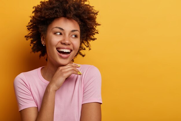 Portrait de jeune femme afro-américaine insouciante positive a un sourire radieux, touche doucement le menton et regarde ailleurs, ravie de remarquer quelque chose d'attrayant, porte des vêtements décontractés, a une beauté naturelle