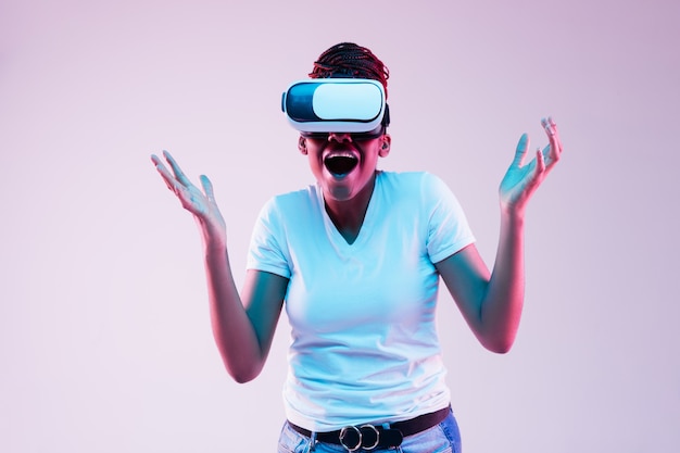 Portrait de jeune femme afro-américaine jouant dans des lunettes VR en néon sur fond dégradé