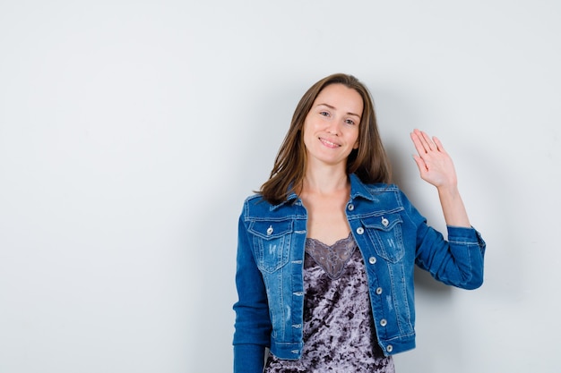 Portrait de jeune femme agitant la main pour saluer en blouse, veste en jean et jolie vue de face