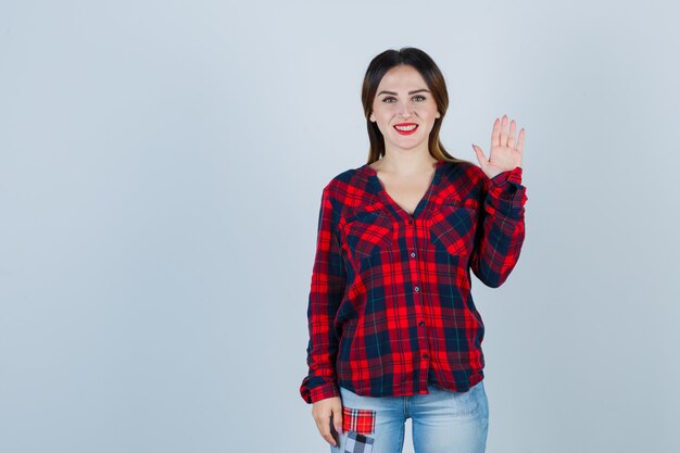 Portrait de jeune femme agitant la main pour saluer en chemise à carreaux, jeans et à la vue de face joyeuse