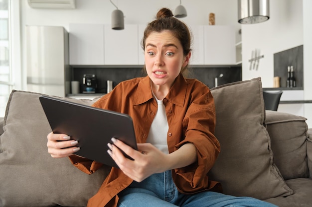 Photo gratuite portrait d'une jeune femme anxieuse et stressée qui a l'air effrayée devant une tablette numérique en train de regarder l'écran assise