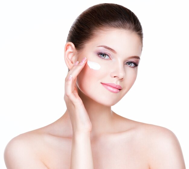 Portrait de jeune femme appliquant de la crème sur son joli visage - sur un mur blanc.