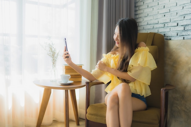 Portrait jeune femme asiatique à l'aide de téléphone portable avec une tasse de café et lire le livre s'asseoir sur une chaise dans le salon