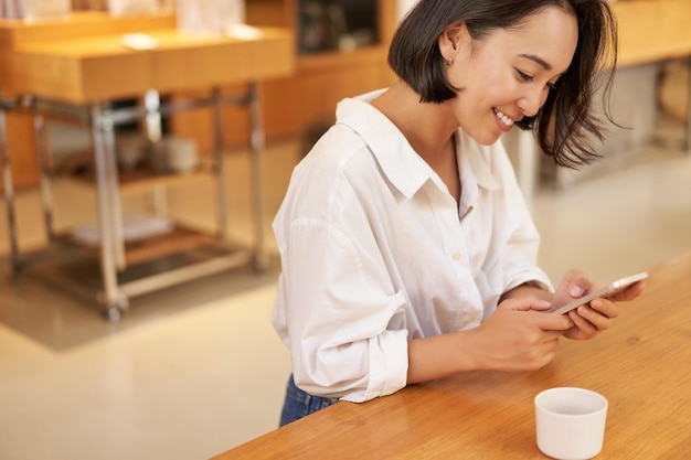 Portrait de jeune femme asiatique assise dans un café tenant un smartphone discutant et envoyant des messages tout en buvant