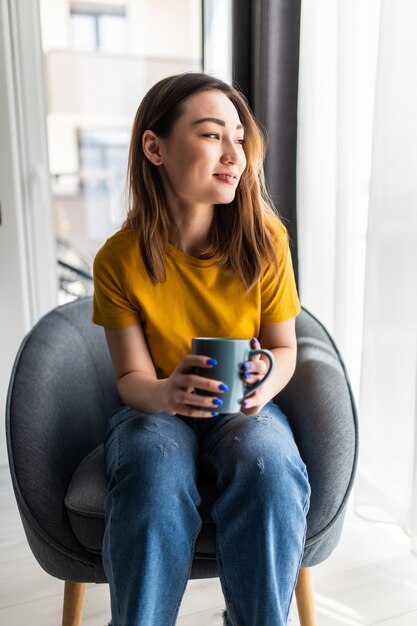 Portrait jeune femme asiatique assise sur un fauteuil avec une tasse de café à l'intérieur du salon