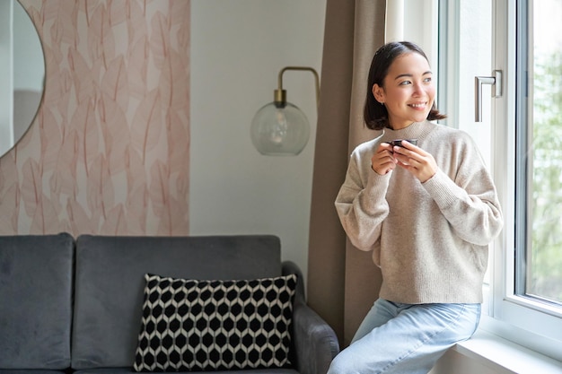 Portrait de jeune femme asiatique assise près de la fenêtre et regardant dehors en buvant du café chaud d'espre