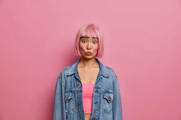 Portrait de jeune femme asiatique aux cheveux bob rose à la mode, a surpris l'expression inquiète, entend quelque chose d'excitant, vêtue d'une veste en jean surdimensionnée, pose à l'intérieur. Fille orientale à la mode