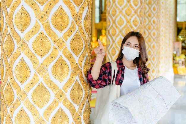 Portrait Jeune femme asiatique routard dans un masque debout et tenir une carte papier à la main dans un beau temple thaïlandais, elle pointe du doigt et attend avec impatience