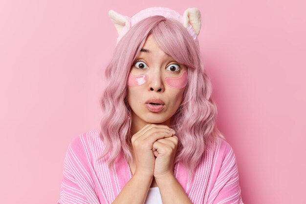 Portrait d'une jeune femme asiatique surprise garde les mains sous le menton fixe les yeux sur écoute applique des patchs d'hydrogel sous les yeux pour le traitement de la peau porte un bandeau isolé sur fond rose. Temps de beauté