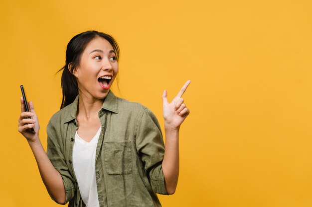 Portrait d'une jeune femme asiatique utilisant un téléphone portable avec une expression joyeuse, montre quelque chose d'étonnant dans un espace vide dans des vêtements décontractés et se tient isolé sur un mur jaune. Concept d'expression faciale.