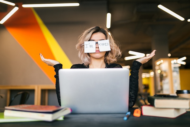 Portrait de jeune femme assez fatiguée avec des autocollants en papier sur des verres assis à table en chemise noire travaillant sur un ordinateur portable dans un bureau de co-working, drôle d'expression, émotion perplexe, problème