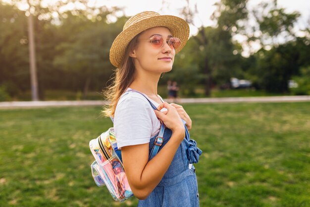 Portrait de jeune femme assez souriante en chapeau de paille et lunettes de soleil roses marchant dans le parc, style de mode estivale, tenue hipster colorée