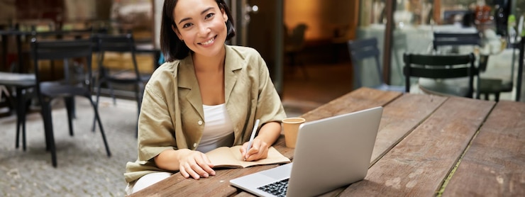 Photo gratuit portrait d'une jeune femme assise dans un café avec un ordinateur portable prenant des notes souriant à la caméra travaillant après