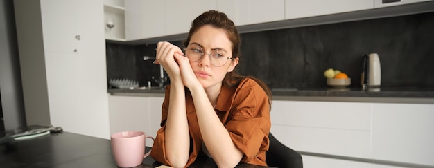 Portrait d'une jeune femme au visage troublé portant des lunettes assise dans la cuisine avec des matériaux de travail sur