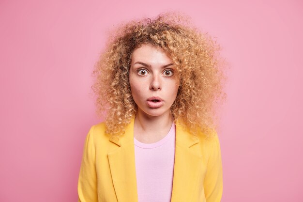Portrait d'une jeune femme aux cheveux bouclés choquée et confuse qui a peur ne peut pas croire que ses yeux vêtus d'une veste jaune isolée sur un mur rose ont surpris l'expression du visage reste sans voix