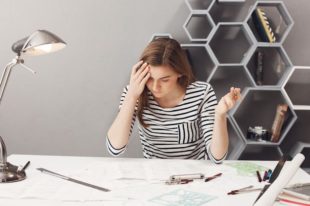 Portrait de jeune femme aux cheveux noirs beau designer freelance en chemise décontractée rayée, écartant les mains, étant frustré, remarquant une grosse erreur dans les calculs financiers