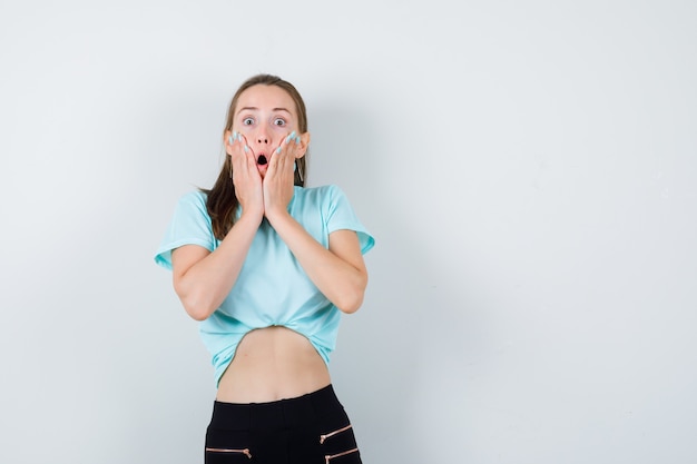 Portrait de jeune femme belle avec les mains sur les joues en t-shirt, pantalon et à la vue de face effrayée