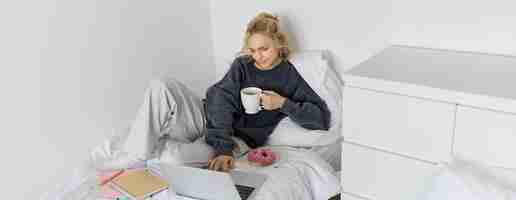 Photo gratuite portrait d'une jeune femme blonde belle se sentant paresseuse à la maison allongée dans le lit avec une tasse de donut de thé