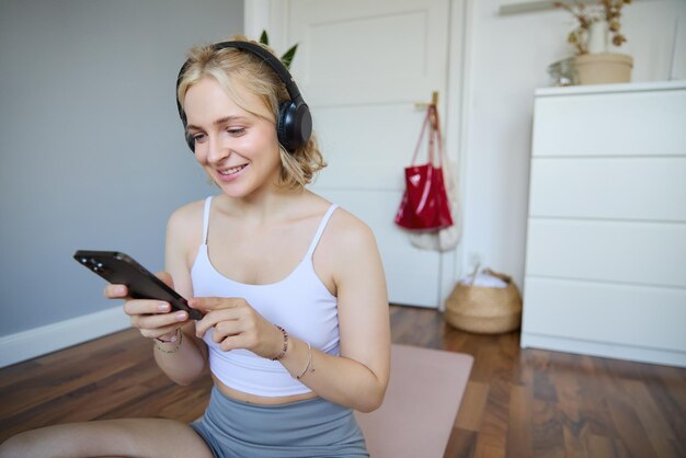 Portrait d'une jeune femme blonde avec des écouteurs qui allume l'application d'entraînement au yoga sur son smartphone en choisissant de la musique