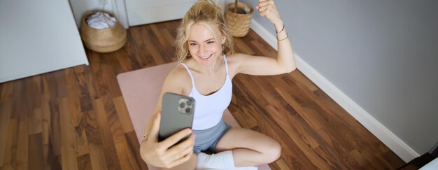 Portrait d'une jeune femme blonde en entraînement physique assise sur un tapis de yoga prenant des selfies pendant l'entraînement à