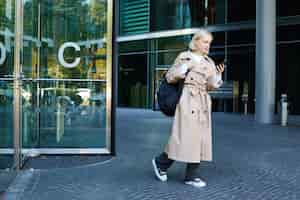 Photo gratuite portrait d'une jeune femme blonde marchant sur un étudiant de la rue avec un sac à dos regardant son téléphone portable