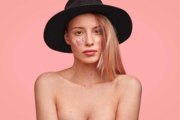 Portrait de jeune femme blonde avec des paillettes sur le visage et un chapeau élégant