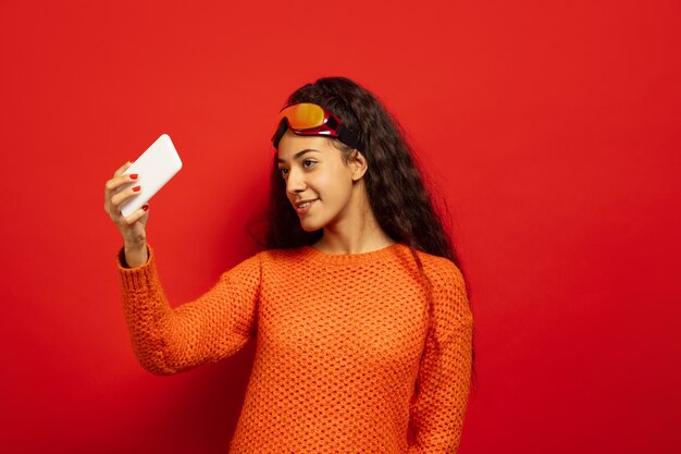 Portrait de jeune femme brune afro-américaine en masque de ski sur fond de studio rouge. Concept d'émotions humaines, expression faciale, ventes, publicité, sports d'hiver et vacances. Boit du thé, du café.