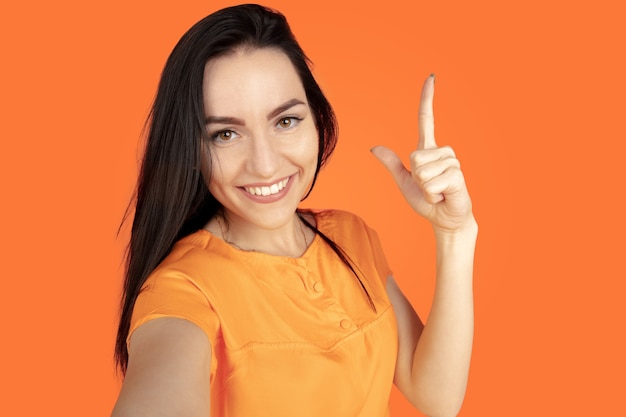 Portrait de jeune femme caucasienne sur fond de studio orange. Beau modèle femme brune en chemise. Concept d'émotions humaines, expression faciale, ventes, publicité. Copyspace. Pointant, montrant, souriant.