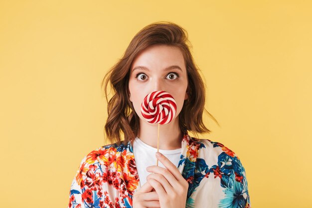 Portrait de jeune femme en chemise colorée debout et couvrant ses lèvres avec des bonbons sucette tout en regardant étonnamment à huis clos sur fond rose