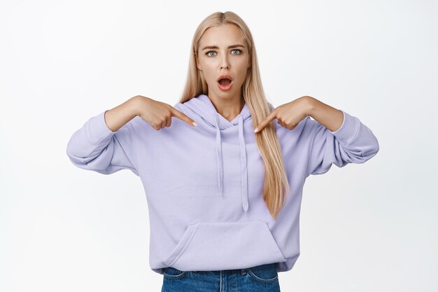 Portrait d'une jeune femme choquée pointant les doigts vers elle-même avec une expression de visage sans voix debout dans un sweat à capuche sur fond blanc