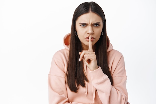 Portrait d'une jeune femme en colère et autoritaire fronçant les sourcils, se taisant à l'avant avec un signe tabou chut sur les lèvres, dites de se taire, grondant une personne bruyante, debout contre un mur blanc