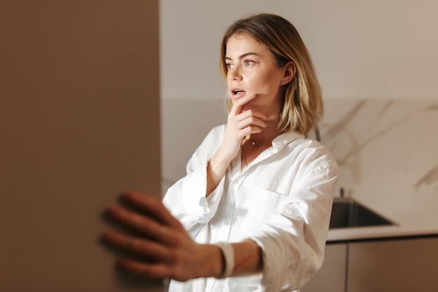 Photo gratuite portrait de jeune femme debout dans la cuisine la nuit et regardant pensivement dans un réfrigérateur ouvert à la maison