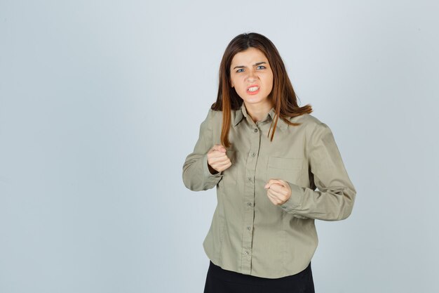 Portrait de jeune femme debout dans une pose de combat, serrant les dents en chemise, jupe et regardant la vue de face en colère