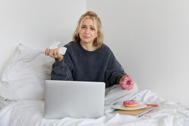 Photo gratuite portrait d'une jeune femme déprimée et déçue pleurant assise sur le lit avec un ordinateur portable mangeant de la nourriture réconfortante