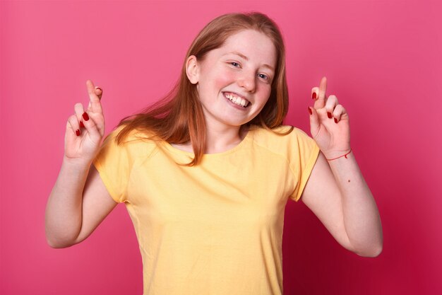 Portrait de jeune femme désireuse en t-shirt jaune décontracté, aux cheveux bruns, croisant les doigts, espère avant un événement important