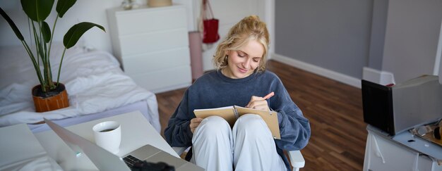 Portrait d'une jeune femme écrivant dans un journal, prenant des notes dans un cahier, assise sur une chaise dans une pièce.