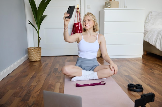 Photo gratuite portrait d'une jeune femme élégante assise sur un tapis en caoutchouc sur le sol à la maison prenant un selfie en faisant