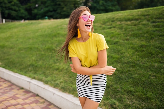 Portrait de jeune femme élégante et séduisante posant dans le parc de la ville, souriant humeur joyeuse, positif, portant haut jaune, mini jupe rayée, sac à main, lunettes de soleil roses, tendance de la mode de style d'été