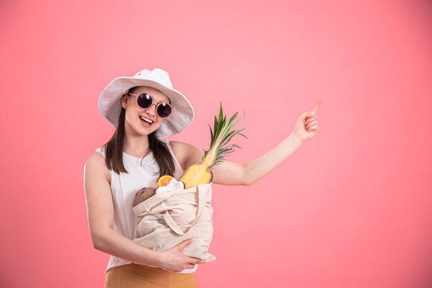 Portrait d'une jeune femme élégante vêtue de vêtements d'été avec un chapeau et des lunettes de soleil, tenant un sac d'éco-fruits, sur rose isolé.