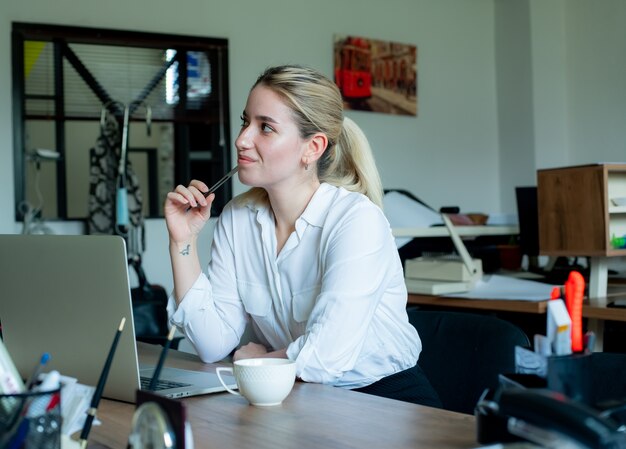 Portrait de jeune femme employée de bureau assis au bureau à l'aide d'un ordinateur portable à la pensée de côté avec une expression pensive travaillant au bureau