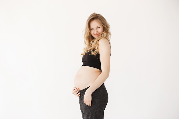 Portrait de jeune femme enceinte joyeuse aux cheveux blonds souriant, tenant le ventre avec la main, regardant à huis clos avec l'expression du visage heureux et satisfait. Beauté et santé