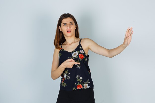 Portrait de jeune femme essayant de se bloquer avec la main, pointant de côté en chemisier, jupe et à la vue de face effrayée